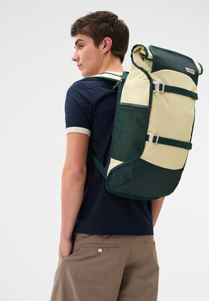 Young man in navy shirt and beige shorts wearing a large beige and dark green backpack, standing with one hand in his pocket, facing sideways.
