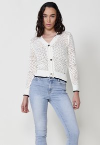 Cárdigan blanco de punto con un patrón de encaje, cuello en V, cierre de botones negros y puños de canalé negros. Combinado con jeans skinny de color azul claro.