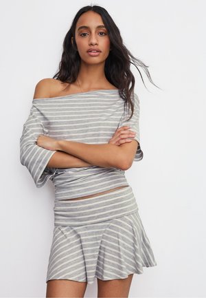 Woman with long dark hair wears gray and white striped off-shoulder top and matching skirt, standing with arms crossed against white background.