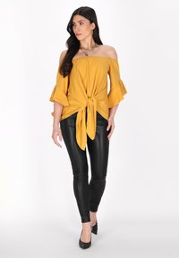 Mouwloze gele blouse met uitlopende mouwen, geknoopte voorkant, gecombineerd met zwarte strakke broek en zwarte hakken.