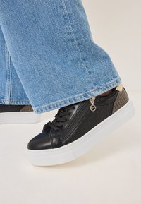 Sneakers in pelle nera con lato decorato, suola platform bianca, lacci e dettaglio con zip. Abbinati a jeans a gamba larga di un blu chiaro.