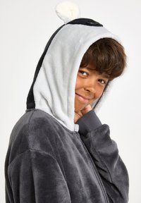 Hoodie en peluche gris avec des oreilles noires et blanches ; texture douce, coupe oversize, fermeture éclair sur le devant et poignets contrastés. Idéal pour le confort et la chaleur.
