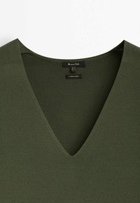Top col en V vert olive en tissu texturé. Présente un col plat et des détails de couture minimaux. L'étiquette est visible au centre.