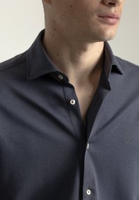 Camisa azul marino con botones, confeccionada en tela suave, que presenta un cuello clásico, botones delanteros visibles y un sutil detalle de logotipo en el pecho.
