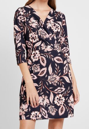 Nainen pukeutunut laivastonsiniseen kietaisumekkoon, jossa on vaaleanpunaisia ja valkoisia kukka- ja lehtikuvioita, kolmen neljänneksen hihat ja V-aukko.