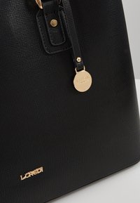 Gros plan sur un sac à main noir texturé avec logo en métal doré et une étiquette décorative ronde dorée accrochée à la sangle.