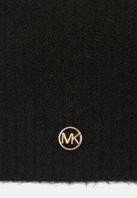 Czarna dzianinowa czapka z miękką teksturą i prążkowanym wzorem, ozdobiona złotym okrągłym logo z napisem "MK" na środku.