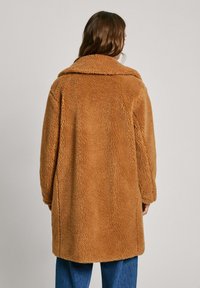 Manteau long, en brown, moelleux, fabriqué dans un tissu doux et texturé, avec un large col, une coupe oversize et sans fermetures visibles.