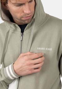 Sudadera con capucha y cremallera en verde claro con detalles en blanco, con bolsillo tipo canguro y la marca "AMARO JEANS" en el pecho. Textura de tela suave.