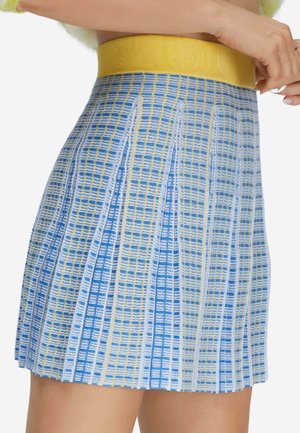 Gestrickter Rock mit blauem, weißem und gelbem Karomuster, breitem gelbem Bund, getragen von einer Person mit verschränkten Armen vor der Brust.