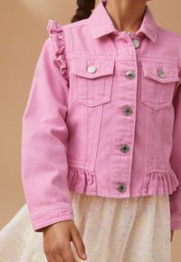 Rosa Jeansjacke mit gerafften Schultern, silbernen Druckknöpfen, zwei vorderen Taschen und einem Volant am Saum, getragen über einem hellen gemusterten Kleid.