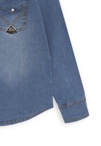 Camicia in denim azzurro chiaro con maniche lunghe, colletto a punta e un accento metallico triangolare con dettagli in catena sulla parte anteriore.