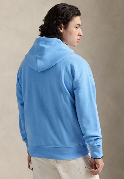 Gaiši zils fleeces džemperis ar kapuci, kas regulējams ar auklu, ribotu apdari aprocēs un apakšmalā, kā arī mīkstu tekstūru. Skats no aizmugures ar minimālu detaļu daudzumu.