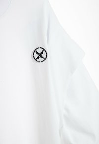 Camiseta blanca de algodón, con un logo bordado negro en el hombro izquierdo. Mangas cortas y un diseño sencillo.