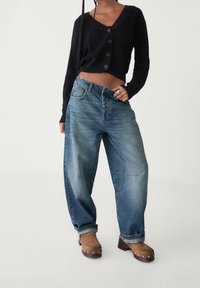 Zwarte geribbelde korte cardigan met knoopsluiting, gecombineerd met lichtblauwe baggy jeans en bruine klompen met zilveren studs.