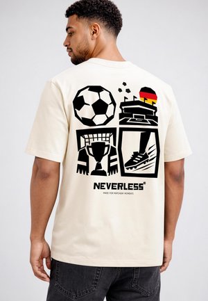 Mann trägt cremefarbenes T-Shirt mit schwarzen Fußball-, Pokal-, Stollenschuh-, Stadion- und Deutschlandflaggen-Grafiken auf dem Rücken, beschriftet mit "NEVERLESS."
