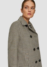 Villakangasta tehty kaksivärinen takki musta-valkoisessa houndstooth-kuosissa, jossa on kauluskäänteet, kaksiriviset napit ja sivutaskut.