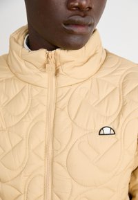 Beige vadderad jacka med hög krage, med dragkedjestängning och en liten broderad logotyp på bröstet. Slät textur och subtil sömnad.