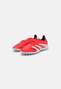 Tacles de football synthétiques rouges avec une tige texturée, des accents noirs et trois rayures blanches ; conçus pour une traction optimale avec une semelle extérieure moulée.