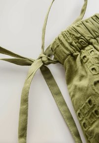 Gros plan d’un tissu vert olive avec un nœud noué, un froncement élastiqué et un détail de broderie découpée décorative.