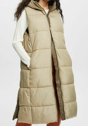 Femme portant une longue doudoune sans manches beige matelassée avec capuche et un haut blanc à manches longues côtelées, mains dans les poches de la doudoune.