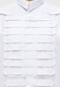 Μπροστινή όψη λευκού πουκαμίσου με οριζόντιες λουρίδες με κουμπιά, τοποθετημένες σε παράλληλες σειρές διαγωνίως στο στήθος και τους ώμους.