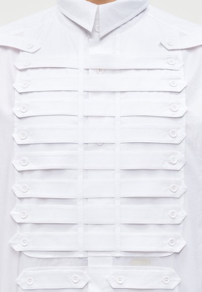 Przód białej koszuli z poziomymi paskami zapinanymi na guziki, ułożonymi równolegle w rzędach na klatce piersiowej i ramionach.