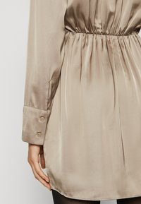 Camisa de satén beige con cintura elástica, mangas largas y puños abotonados. Textura suave, aberturas laterales en el dobladillo y detalles con pliegues.