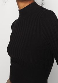 Haut en maille côtelée noire avec un col montant, présentant un design texturé et une silhouette ajustée, mettant en valeur des rayures verticales et un tissu lisse.