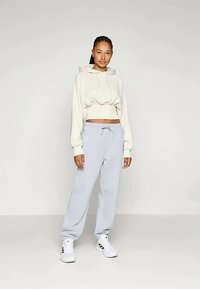 Sudadera crema recortada con cordón, combinada con pantalones de chándal gris claro. El modelo lleva zapatillas blancas con acentos negros. Texturas suaves.