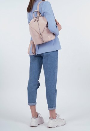 Mochila de color rosa claro con exterior texturizado, correas curvas dobles y cremallera metálica. Se lleva en la espalda, combinada con vaqueros azules y zapatillas claras.