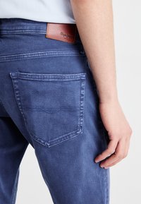 Temno modri jeans iz denima z gladko teksturo, ki imajo logotip na pasu in en zadnji žep z obšitimi detajli.