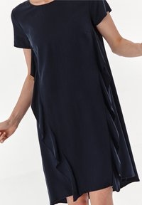 Navyblaues Kleid mit kurzen Ärmeln, das einen lockeren Schnitt und asymmetrische Rüschendetails an den Seiten aufweist. Glattes, leichtes Material.