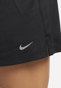 Mustat urheilushortsit, joissa on teksturoitu kangas, pieni hopeinen Nike-logo alareunassa ja lievä halkio helmassa.