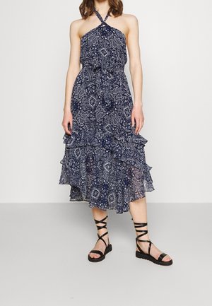 Vrouw draagt een mouwloze marineblauwe halterjurk met witte paisleypatronen en zwarte platte sandalen met bandjes, staand tegen een effen achtergrond.