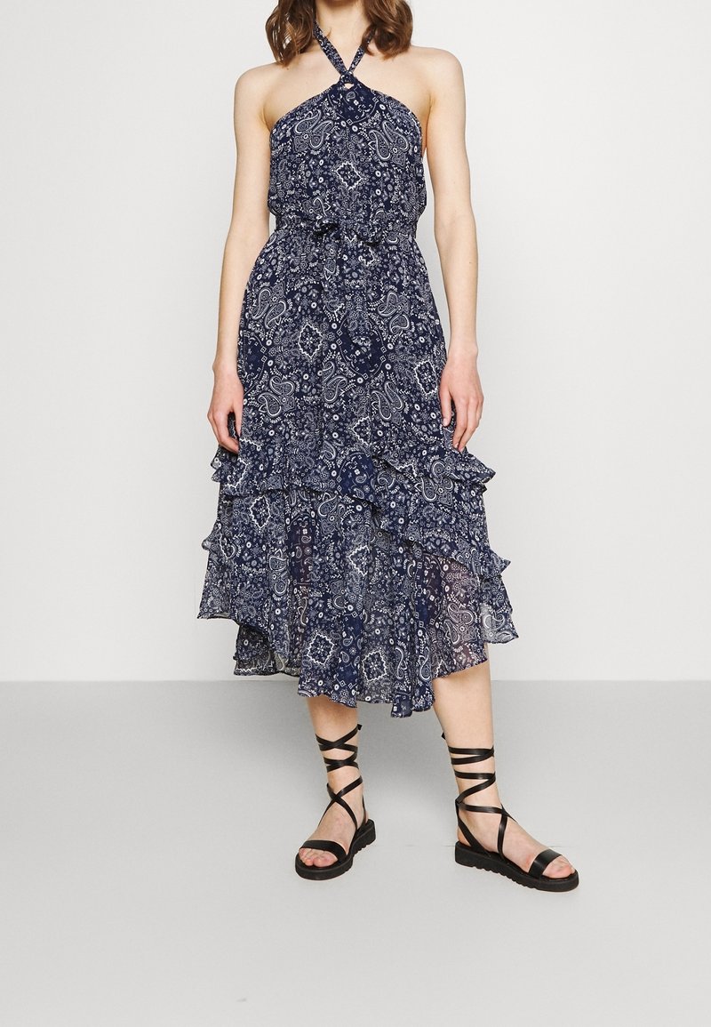 Femme portant une robe dos nu bleu marine avec des motifs paisley blancs et des sandales plates noires à lanières, debout devant un fond uni.