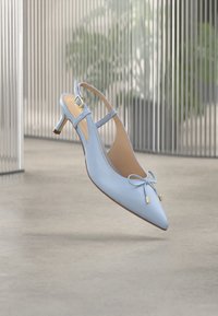 Világoskék bőr hátul pántos magassarkú hegyes orral, apró masnival elöl és arany csattal, semleges beltéri háttér előtt.