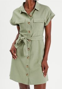 Robe chemise verte olive avec des manches courtes, boutons sur le devant, deux poches poitrine et une ceinture assortie à la taille. Texture de tissu lisse.