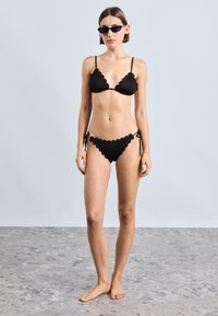 Black bikini made of smooth fabric featuring scalloped edges, a triangular top with thin straps, and a hip-tie bottom, paired with black sunglasses.