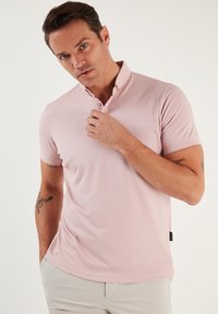 Világos rózsaszín polo póló, slim fit fazonnal, rövid ujjal, és két gombbal ellátott gallérral. Sima anyag textura és diszkrét varrási részletek.