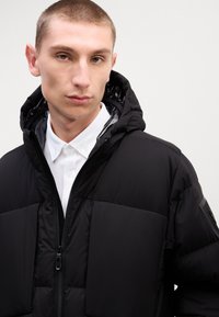 Jachetă puffer neagră cu glugă, dotată cu închidere cu fermoar, textură matlasată și căptușeală interioară lucioasă. Purtați peste o cămașă albă cu guler.
