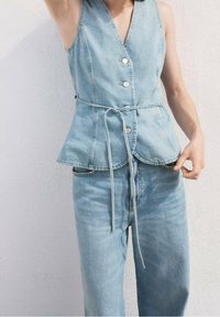 Lyseblå denimvest med peplumdesign, der har en v-hals, knappedetalje og snøretalsnørre i taljen, kombineret med matchende jeans.