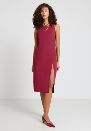 Woman with curly hair wearing sleeveless maroon dress with thigh slit and matching heels, standing against plain white background.