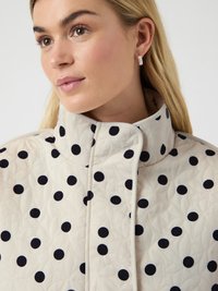 Ξανθιά γυναίκα που φοράει μπεζ μπουφάν με επένδυση, ψηλό γιακά με μεγάλα μαύρα πουά και ασημένια σκουλαρίκια κρίκους.