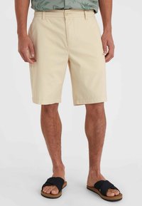 Beige Baumwollshorts mit einem Knopfverschluss vorne, Gürtelschlaufen und seitlichen Taschen. Getragen mit schwarzen Riemensandalen. Glatte Textur, knielang.