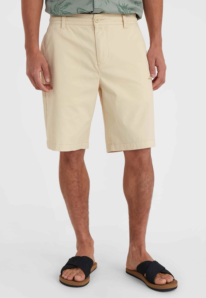 Beige Baumwollshorts mit einem Knopfverschluss vorne, Gürtelschlaufen und seitlichen Taschen. Getragen mit schwarzen Riemensandalen. Glatte Textur, knielang.