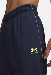 Navy athletic shorts with elastic waistband, featuring a neon yellow side accent and Under Armour logo on the lower leg. Smooth texture.