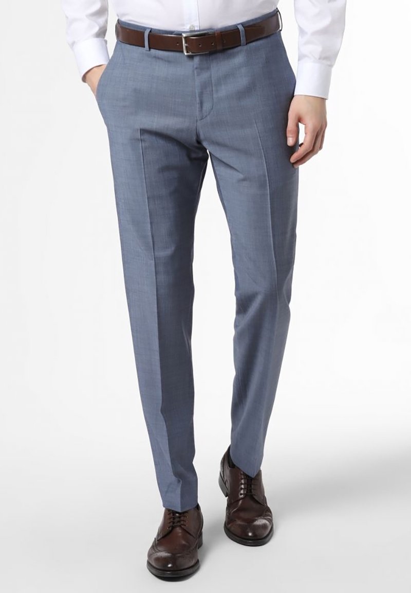 Homme portant un pantalon de costume bleu clair, chemise blanche rentrée dans le pantalon, ceinture en cuir marron et chaussures de ville marron foncé, debout avec une main dans la poche.