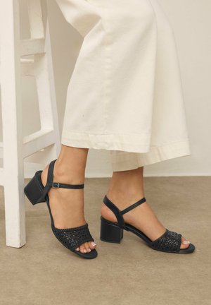 Pieds portant des sandales à talons tressées noires avec des brides à la cheville, associés à un pantalon large blanc, debout à côté d'une chaise blanche sur un sol beige.