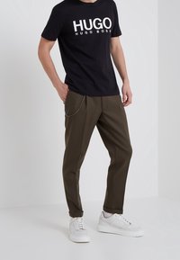 Zwart katoenen T-shirt met "HUGO" in witte letters, gecombineerd met donkerolijfgroene broek met kettingaccent en witte sneakers.